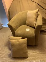 Front angled view of beige faux suede swiveling armchair with one cushion on the seat and two cushions in front on carpeted floor.