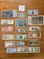 Photo of 18 assorted foreign currency bills arranged on a wooden surface, showing different denominations, colors, and designs.