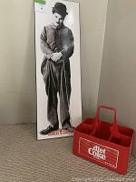 Full view of tall, narrow Charlie Chaplin black and white art picture standing upright next to a red Diet Coke bottle holder on floor.