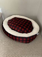 Front/angled view of oval pet bed with red and black Canadian plaid cushion surrounded by white sherpa border.