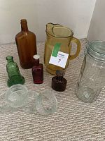 Full view showing all nine mixed colour glassware items on carpet: yellow glass pitcher, amber G & W bottle, green Wheaton Liberty Bell bottle, red and green smaller bottles, tall clear locking glass container, two small clear glass vases.