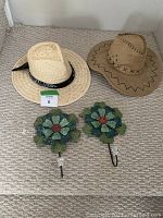 Two sun hats and two metal flower wall hooks on carpet. Hats include a woven straw fedora with black bandanna and a suede-like hat with decorative stitching. Hooks are green metal flowers with red centers.