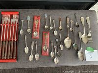 Photo showing 6-piece fondue fork set with wooden handles in a red box, and a variety of 21 collectible spoons with diverse designs laid out on a table.