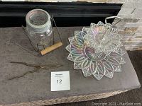 Front view of antique metal pickling lifter with Mason jar, metal pickling tongs, and vintage iridescent two-tier glass dessert tray on a brown bench.