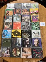 28 assorted CDs arranged flat on a wooden table, showing various artists and albums, displaying album covers and titles.