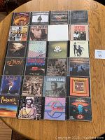 Full layout showing all 24 mixed rock CDs lying flat on a wood table, arranged in rows. Covers include classic rock and modern rock artists.