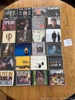 Top-down photo of 24 rock music CDs arranged on wooden table showing cover art for various artists including Pearl Jam, Johnny Cash, Uncle Kracker, Bob Dylan, Bruce Springsteen, and more.