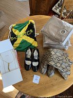 Overview photo showing all lot items including boots in a Jamaica-themed bag, black patent Mary Jane shoes, beige curtains in plastic packaging, and the gold colored necklace in box.