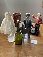 Photo shows four wine bottle holders on a wooden surface: a cream-colored wine burka cover, large hand carved wood upright holder, metal guitar player figure, and "Carl the Waiter" figurine with broken fingers.