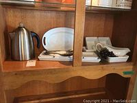 Wide view of assorted white ceramic dishes, a stainless steel coffee percolator, and additional items on wooden shelving