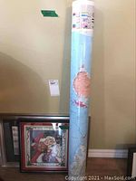 Rolled laminated world map shown standing vertically against wall beside framed artworks on floor.