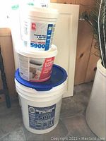 Stack of three buckets: 5-gallon waterproof rubber membrane bucket with blue lid at the bottom, partially full 4-liter E-z Roll Stucco bucket in the middle, smaller unopened bucket of ceramic tile adhesive on top, next to a white kitchen cupboard door.