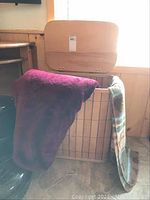Laundry hamper made of light wood with hinged lid and black strip accents with blankets draped over side.