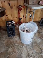 Overall view of wine making supplies grouped together including red bottle corker, bucket with agitator rod, and other tools