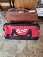 Photo showing the Milwaukee branded red and black duffel bag in front of a brown vintage suitcase.