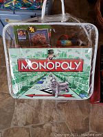 Monopoly board game in clear carrying case with visible game pieces and box cover