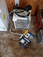 Photo showing white portable heater with attached power cord, chrome faucet with flexible hose, and boxed Weiser door knob set on floor.