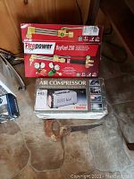 Photo showing the boxed Firepower OxyFuel 250 oxy-acetylene torch and the boxed Motomaster air compressor with a small buck saw on the floor.