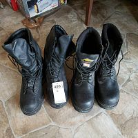 Two pairs of black work boots placed side by side on a stone-tile floor, both showing signs of wear, one pair includes a tag.