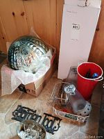 Large decorative gazing ball in bubble wrap, boxed garden arbour, mosquito lanterns, red metal vase, chandelier crystals in box, garden tool holder on floor