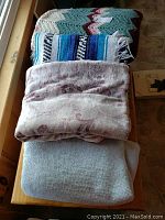 Four blankets stacked on a wooden bench near a window. The top blanket is a crocheted zig zag pattern afghan in cream, sage green, burgundy, and light pink colors. Underneath is a woven blue striped blanket with white, black, and turquoise colors and fringed ends. Below are two soft textured blankets, one light pink and one light blue.