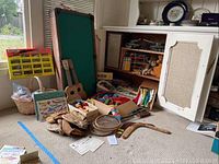 Wide view of assorted vintage children's toys arranged near a cabinet including ukuleles, baseball gloves, wooden blocks, badminton rackets, boomerangs, and pool table.
