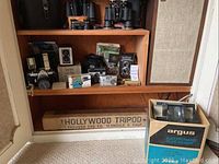 Overview of wooden shelving displaying multiple cameras, binoculars on top shelf, tripod case and Argus slide viewer on lower shelf