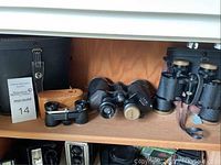 Three vintage binoculars including Swift Stadium and Sunset brand shown on wooden shelf with leather and black cases.