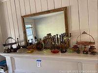 Gold colored mirror and various decorative items on shelf including brass candlesticks, colored glass dishes, metal trays with handles, and gold frame without backing