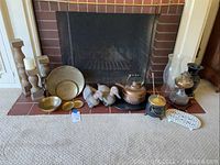 Overview of lot contents on fireplace hearth: wood candlesticks, brass bowls and platter, duck decoys, copper kettle, small pot, oil lamp, glass vase, trivet, poker