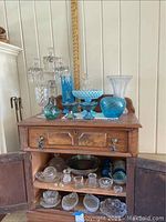 Wide view of various blue and clear glass items on wooden chest, including candelabra, vases, bowls, and glasses.