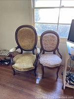 Two Victorian style chairs side by side by a window on wooden floor. Larger chair with gold velvet upholstery, smaller chair with floral upholstery and casters.