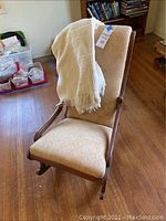 Angled view of small vintage wooden rocking chair with beige upholstery and cream throw blanket draped on back