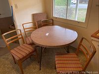 Full view of round vintage Formica table with metal legs and 4 wooden ladder-back chairs with upholstered seats in room corner near window.