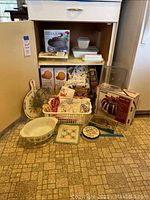 Photo of vintage and newer kitchen items arranged on floor and shelf, showing Pyrex dishes, aprons, baking pans, and cookbooks