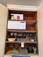 Wood kitchen cabinet shelf displaying assorted glass plates, bowls, serving bowl, glasses, cream and sugar set, and wine glasses in boxes.