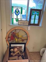 Three stained glass pieces displayed near window, including one round panel and two rectangular panels. Below, a box of assorted framed art and decorative items.