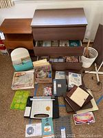 Wide view of scrapbooking supplies including photo albums, glue, cutter, and cards inside dresser drawers and on the floor