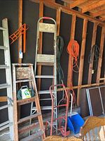 Three ladders of varying sizes leaning against a wood beam garage wall. Two red hand dollies, blue dustpan, and dustbin on the floor. Multiple extension cords hanging on wall.