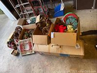Boxes containing assorted holiday decorations including wreaths, bows, and figurines placed on a cardboard box in a garage setting.