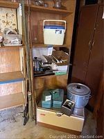 Wide view showing cooler at base, two green metal tackle boxes, stacked cookware on shelf and three fishing rods leaning against shelving.