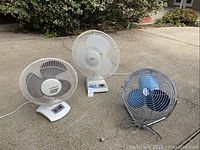 Three electric fans of various sizes and brands on concrete surface outside. From left to right: Cool-Breeze, Aries, and a large blue blade floor fan.
