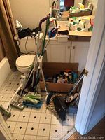 Photo of bathroom floor with multiple brooms, mops, a box of cleaning agents and supplies on the floor near a toilet and sink cabinet.