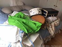 Photo showing multiple items including green fabric storage cover, hat box, luggage and vintage suitcase on bed with floral bedspread.