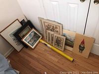 Collection of framed and unframed art prints and rolled art, leaning against door on hardwood floor showing variety of frames and art styles.