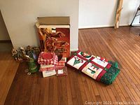 Full view of the lot including Grandeur Noel Nativity figures, box, Christmas quilt folded on floor, picnic accessories and wind chime near a wall on wood floor