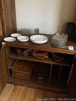 Photo showing wooden serving trays stacked on a bottom shelf and wooden bowls on an upper shelf with glass plates and Corinthian by Sebring plates on top of a wooden stand.