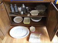 Wide view inside wooden cabinet showing Corning Ware dishes, Pyrex bowl, ceramic turkey platter, chopsticks bag on floor, other small kitchen items