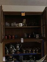 Wooden cabinet with upper and lower shelves displaying metal cups, coffee pots, mugs, and decorative items including owl figure and bonsai tree.