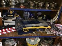 Shelf showing silver trophy cups, Naval Academy pennant, cap, bumper sticker, buttons, and folded cloth items.